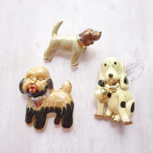 May include: Three enamel dog pins. One is a brown and white dog with a gold collar. One is a black and white dog with a gold collar and a bell. One is a white and black dog with a gold collar.