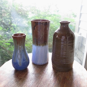 May include: Three ceramic vases in varying sizes and shapes. The vase on the left is blue and brown, the middle vase is brown and light blue, and the vase on the right is brown. The vases are on a wooden table.