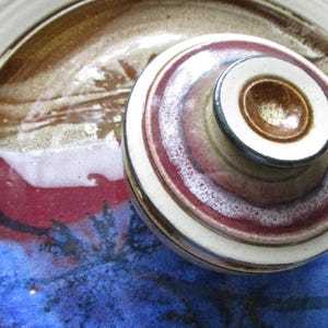 May include: A handmade ceramic pottery set with a lid. The lid and base feature concentric rings of color, including white, red, brown, and blue. The pottery is likely used for food storage or display.