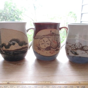 May include: Three ceramic mugs with coastal designs. The mugs feature illustrations of a sailboat, seashells, and a lighthouse. Each mug has a handle and a blue base, with earth-toned and white accents.