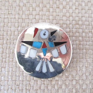 May include: A silver round pin with a Native American design. The design features a stylized bird with turquoise, red, black, and white stones.