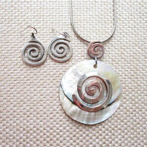 May include: A silver necklace with a large, round, white shell pendant featuring a silver spiral design. The necklace is paired with matching silver earrings with a spiral design.