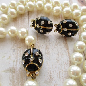 May include: A vintage jewelry set featuring a pearl necklace and matching earrings. The earrings and pendant have a black enamel finish with gold accents and are adorned with sparkling rhinestones. The pearls are a creamy white color.
