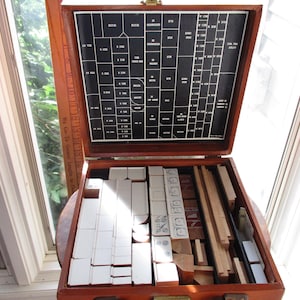 May include: Open wooden box containing various blocks and pieces, likely for a game or educational activity. The box has a black interior with white text and diagrams. A wooden ruler is visible on the side. The box is open, revealing the contents.
