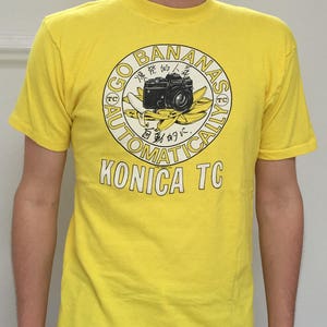 Peut inclure: T-shirt jaune avec un motif rond représentant un appareil photo noir, des bananes et le texte "GO BANANAS AUTOMATICALLY KONICA TC". Le design est de style vintage.