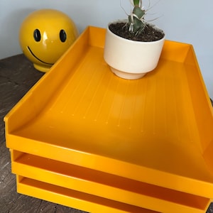 May include: A stack of three bright yellow plastic trays. A small white ceramic pot with a cactus sits on top of the trays. A yellow smiley face ceramic ball is in the background.