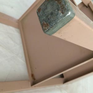 K&ouml;nnte beinhalten: Ein rosa Metalltisch mit einem Metallbein, das an der Spitze eine graue Metallkappe hat. Der Tisch hat ein Regal.
