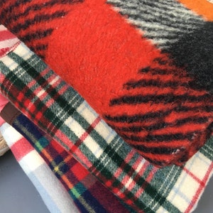 May include: A stack of four blankets in various plaid patterns. The top blanket is red, black, and white with a fuzzy texture. The second blanket is red, green, and white plaid. The third blanket is red, blue, and white plaid. The bottom blanket is blue and white plaid.
