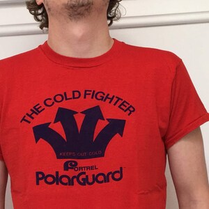 Puede incluir: Camiseta roja con un gráfico azul que dice "The Cold Fighter" con tres flechas apuntando hacia arriba y el texto "Keeps Out Cold" y "PolarGuard" con un logotipo.