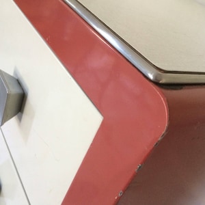 May include: A white dresser with three drawers and red trim. The drawers have silver metal handles. The top of the dresser is a light gray color.