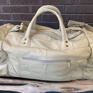 Op de afbeelding: Een lichtbeige leren reistas met twee bovenste handvatten, een ritssluiting en een voorvak met een ritssluiting. De tas heeft een versleten, vintage uitstraling.