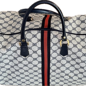 Puede incluir: Una bolsa de lona vintage con un patrón repetido de logotipos dobles-G entrelazados en azul y blanco. La bolsa presenta una franja central roja, detalles en azul marino y herrajes dorados. Tiene un asa superior y un cierre de cremallera.