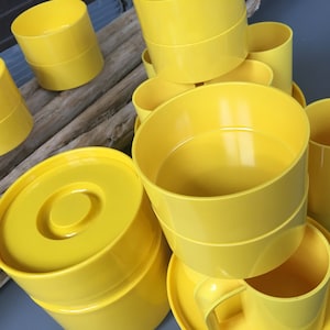 以下が含まれることがあります： 木製の表面に積み重ねて並べられた、黄色いプラスチック製のカップとボウル。カップにはハンドルが付いており、ボウルは丸い形状です。