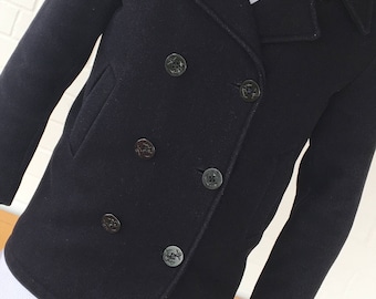 May include: A dark blue peacoat with a double-breasted closure and six black buttons. The coat has a traditional sailor collar and a single pocket on the left side.
