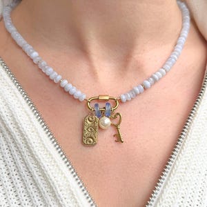 May include: A beaded necklace featuring light blue translucent beads and a gold-toned carabiner clasp. The clasp holds three charms: a rectangular gold pendant with an eye and celestial motifs, a white pearl bead, and a gold-toned skeleton key.