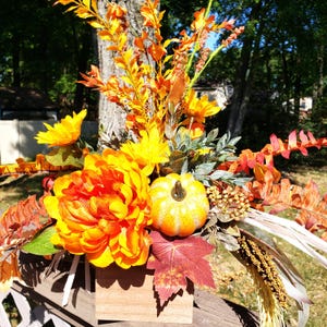 May include: A fall-themed floral arrangement in a wooden box. The arrangement features large orange and yellow flowers, a small pumpkin, and various fall foliage. The background includes a tree and a glimpse of a house.