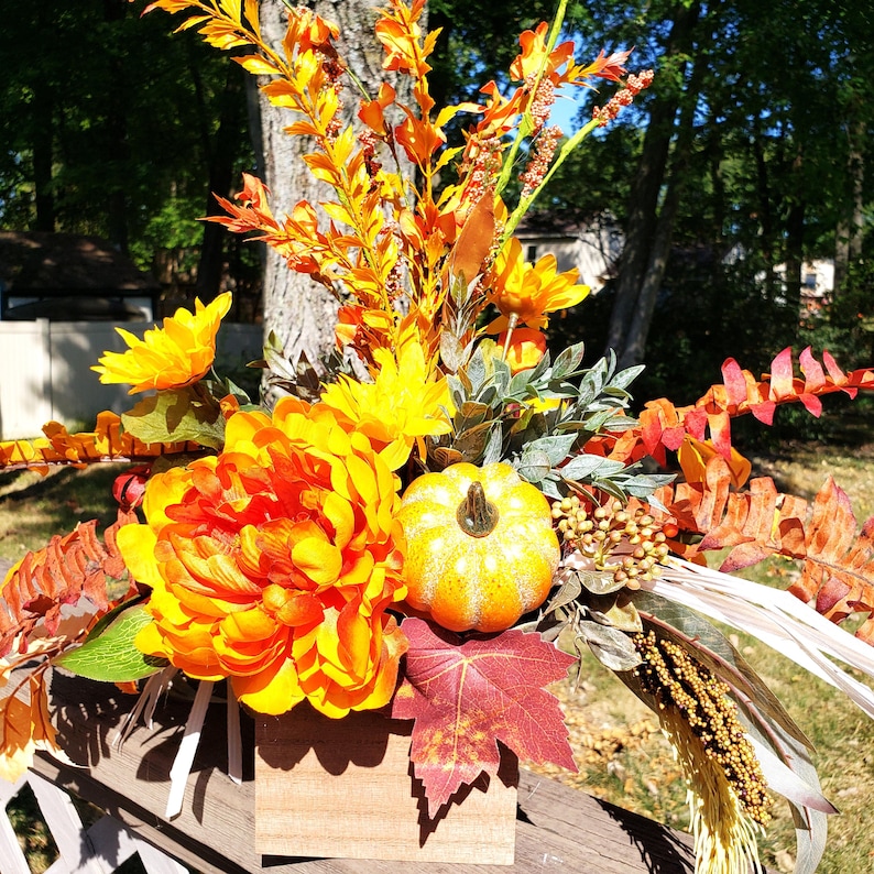 May include: A fall-themed floral arrangement in a wooden box. The arrangement features large orange and yellow flowers, a small pumpkin, and various fall foliage. The background includes a tree and a glimpse of a house.