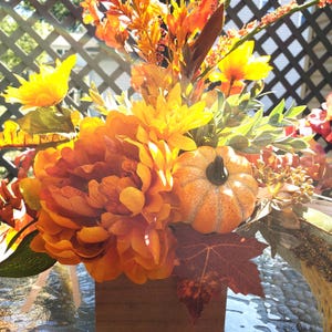 May include: A fall-themed floral arrangement featuring orange and yellow flowers, a small pumpkin, and fall foliage. The arrangement is in a wooden box, perfect for autumn decor.