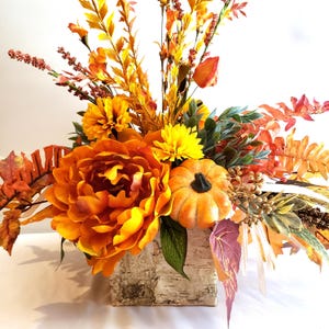 May include: A fall-themed floral arrangement featuring a large orange peony, yellow chrysanthemums, and a small pumpkin. The arrangement includes various orange and yellow foliage, displayed in a birch bark-style container. The overall color palette is warm and autumnal.