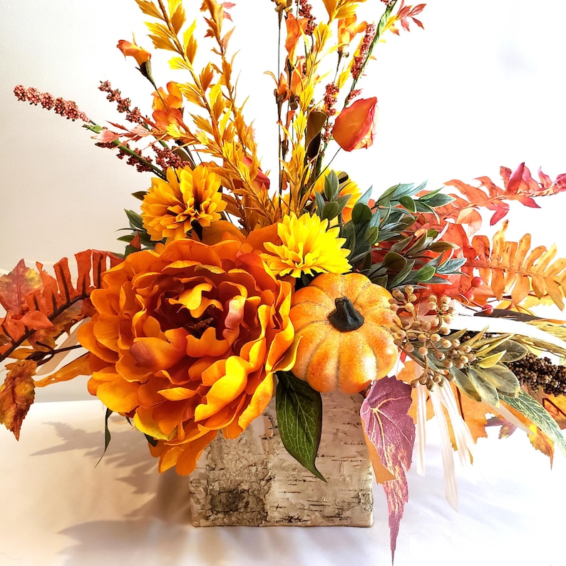 May include: A fall-themed floral arrangement featuring a large orange peony, yellow chrysanthemums, and a small pumpkin. The arrangement includes various orange and yellow foliage, displayed in a birch bark-style container. The overall color palette is warm and autumnal.