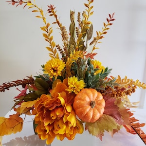 May include: A fall-themed floral arrangement featuring a small orange pumpkin, yellow chrysanthemums, and orange and brown leaves. The arrangement includes various types of foliage and berries, creating a festive and seasonal display. The flowers are arranged in a low, wide container.