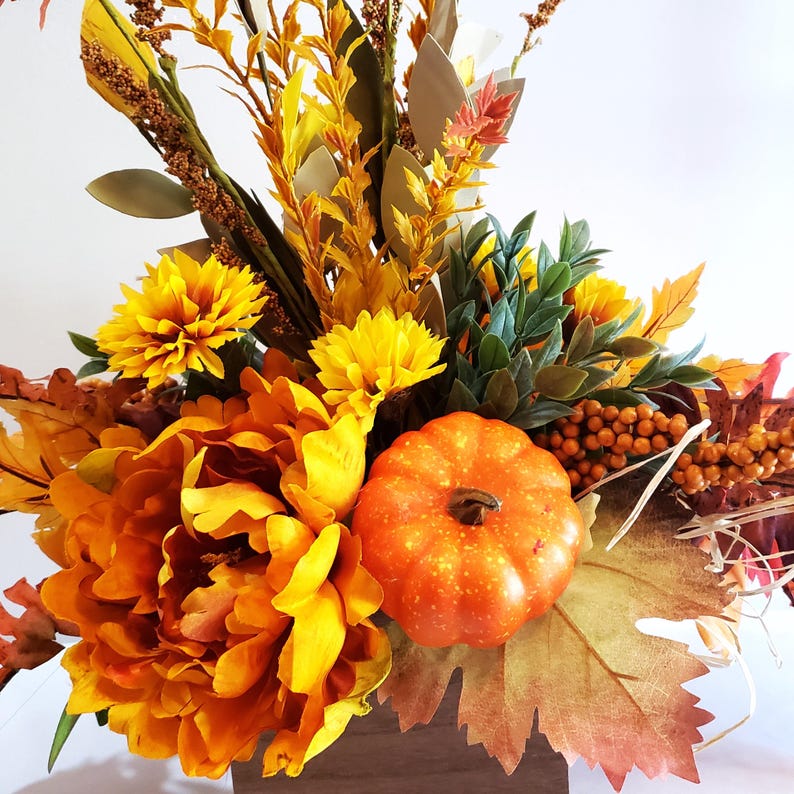 May include: A fall-themed floral arrangement in a wooden box. The arrangement features large orange and yellow flowers, a small orange pumpkin, and various fall foliage. The colors are warm and inviting, perfect for autumn decor.