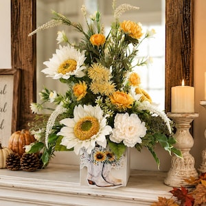 May include: A floral arrangement featuring white sunflowers, yellow chrysanthemums, and green foliage in a white square vase decorated with a graphic of cowboy boots and sunflowers. The arrangement is displayed with two candles and pumpkins.
