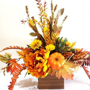 May include: A fall-themed floral arrangement in a wooden box. The arrangement features orange and yellow flowers, including a large peony, chrysanthemums, and a small pumpkin. Various fall foliage and berries complete the display.
