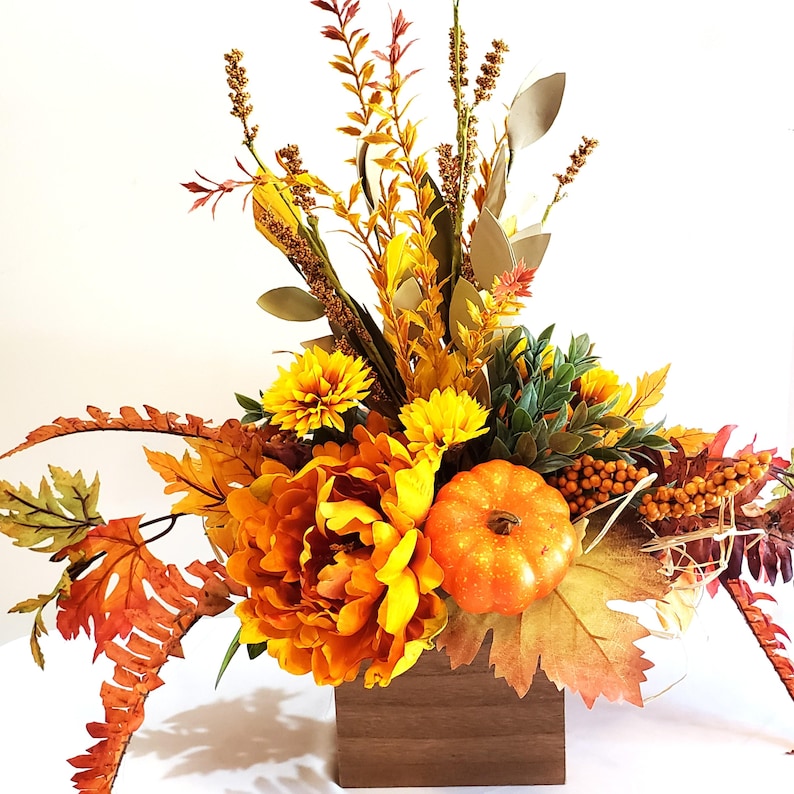 May include: A fall-themed floral arrangement in a wooden box. The arrangement features orange and yellow flowers, including a large peony, chrysanthemums, and a small pumpkin. Various fall foliage and berries complete the display.