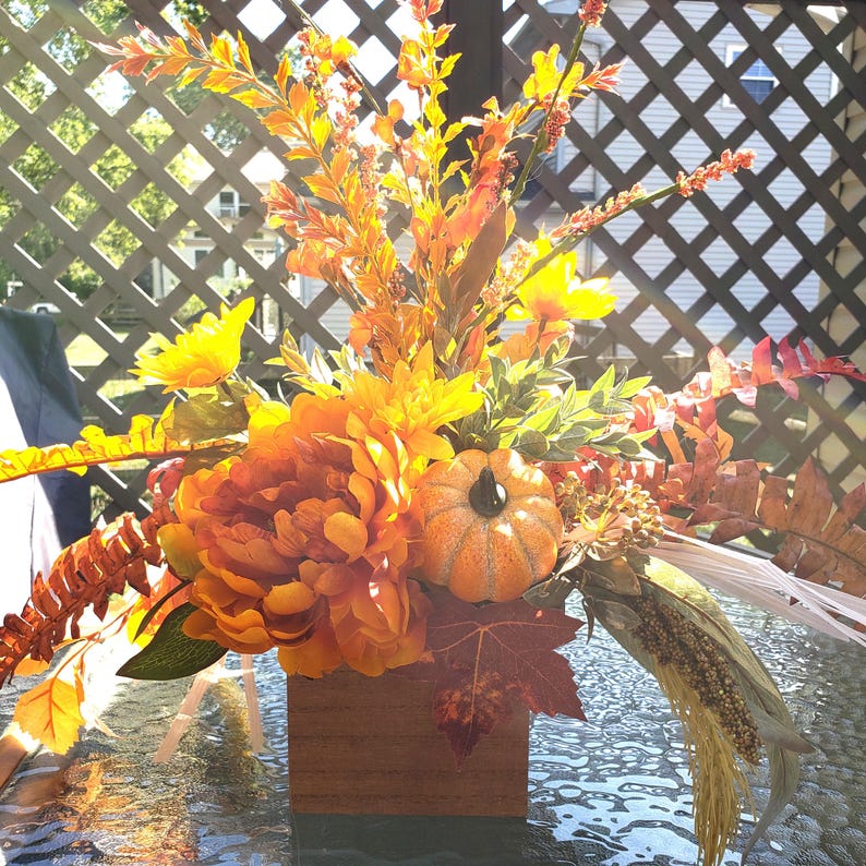 May include: A fall-themed floral arrangement in a wooden box. The arrangement features orange and yellow flowers, greenery, and a small pumpkin. The flowers are arranged in a square wooden box, creating a warm and inviting display.