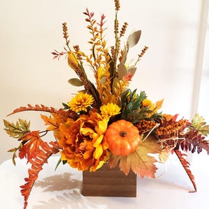 May include: A fall-themed floral arrangement in a wooden box. The arrangement features orange and yellow flowers, a small pumpkin, and various fall foliage. The colors include shades of orange, yellow, green, and brown. The arrangement is set on a white surface.