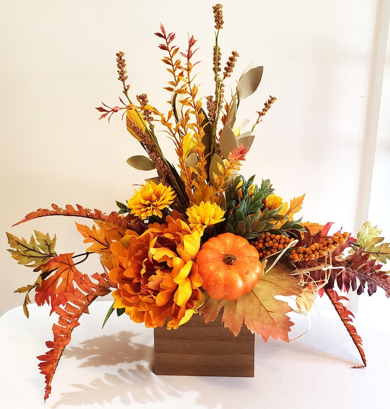May include: A fall-themed floral arrangement in a wooden box. The arrangement features orange and yellow flowers, a small pumpkin, and various fall foliage. The colors include shades of orange, yellow, green, and brown. The arrangement is set on a white surface.