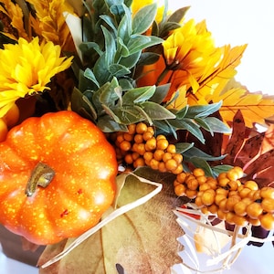 May include: A fall-themed arrangement featuring a small orange pumpkin, yellow flowers, green leaves, and orange berries. The arrangement includes faux fall leaves and is likely used for seasonal home decor.