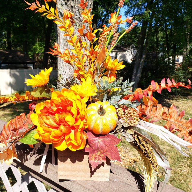 May include: A fall-themed floral arrangement featuring a large orange peony, yellow sunflowers, and a small pumpkin. The arrangement includes various orange and red leaves and is set in a small wooden box. The background shows a tree and a glimpse of a house.