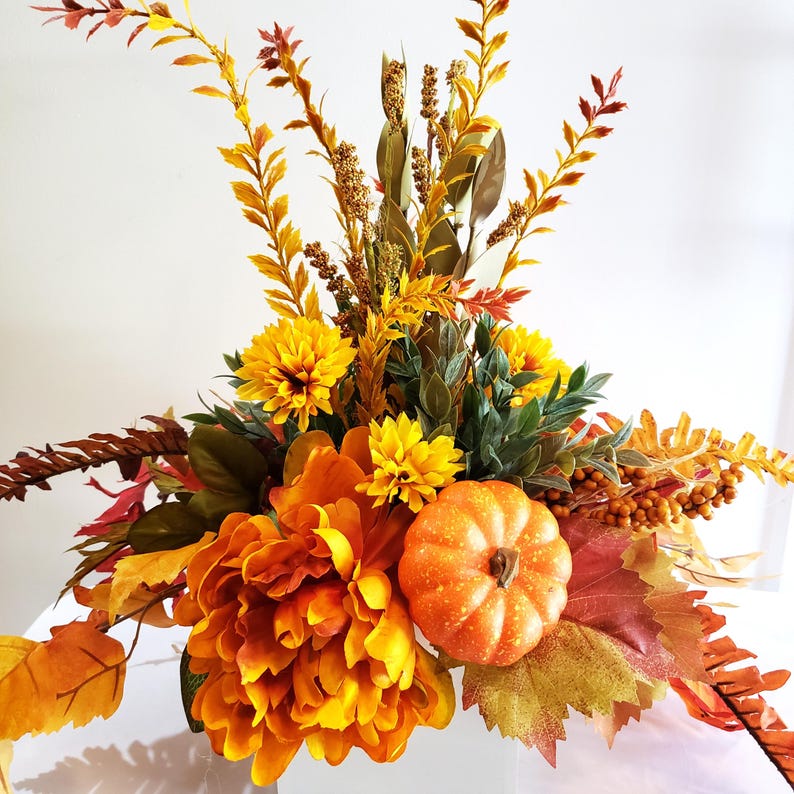 May include: A fall-themed floral arrangement featuring vibrant orange and yellow flowers, decorative leaves, and a small pumpkin. The arrangement includes various textures and shades of autumn colors, creating a festive centerpiece.