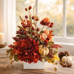 May include: A fall-themed floral arrangement in a white square vase. The arrangement features deep red dahlias, orange roses, and small pumpkins. A lit candle, books, pinecones, and a small white pumpkin are also on the wooden table.