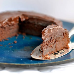 May include: A slice of chocolate cake on a blue plate with a silver cake server. The cake is covered in a rich chocolate frosting.