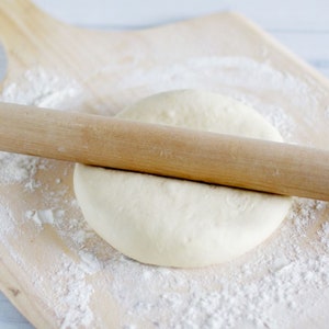 May include: A wooden rolling pin is rolling out a round piece of dough on a wooden cutting board covered in white flour.