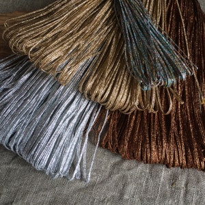 Pode incluir: Uma cole&ccedil;&atilde;o de cord&otilde;es met&aacute;licos em prata, ouro, marrom e azul iridescente. Os cord&otilde;es est&atilde;o dispostos em forma de leque e s&atilde;o feitos de um material brilhante e texturizado.