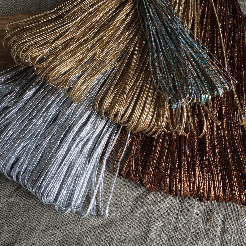 Pode incluir: Uma cole&ccedil;&atilde;o de cord&otilde;es met&aacute;licos em prata, ouro, marrom e azul iridescente. Os cord&otilde;es est&atilde;o dispostos em forma de leque e s&atilde;o feitos de um material brilhante e texturizado.