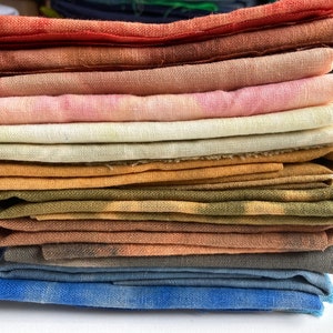 Op de afbeelding: Een stapel van 11 gevouwen vierkante stukken stof in verschillende tinten bruin, oranje, roze, geel, groen en blauw. De stof is van natuurlijk linnen.