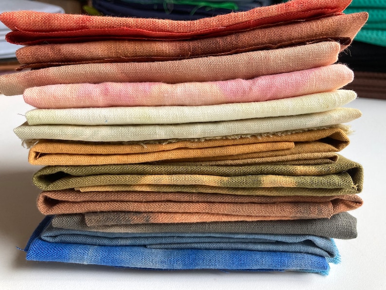 Op de afbeelding: Een stapel van 11 gevouwen vierkante stukken stof in verschillende tinten bruin, oranje, roze, geel, groen en blauw. De stof is van natuurlijk linnen.