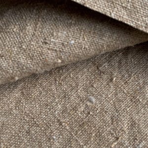 Może przedstawiać: Zbliżenie na teksturowaną tkaninę w kolorze beżowym. Widoczny jest splot, o lekko szorstkim wyglądzie i małych, jasnych płatkach. Tkanina jest złożona, tworząc cienie i głębię.