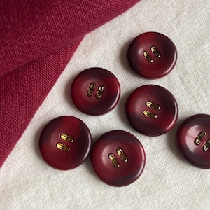 Puede incluir: Seis botones redondos con un efecto veteado rojo oscuro y morado, cada uno con dos agujeros dorados. Un trozo de tela roja doblada está en la esquina superior izquierda, y los botones están sobre una superficie blanca.