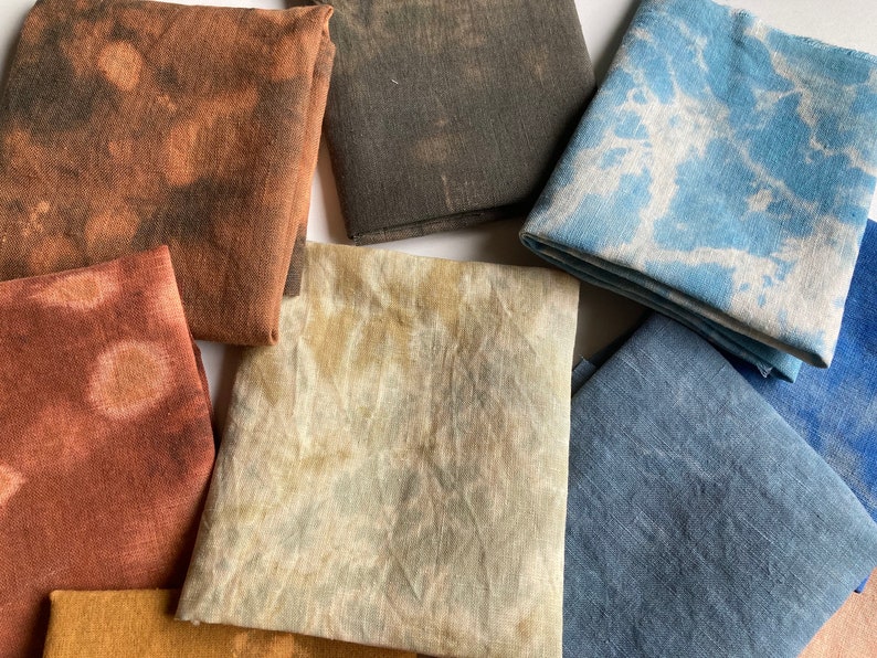 Op de afbeelding: Een verzameling van acht stoffen vierkanten in verschillende tinten bruin, blauw en beige. De stoffen vierkanten hebben een tie-dye effect en zijn in een willekeurig patroon gerangschikt.