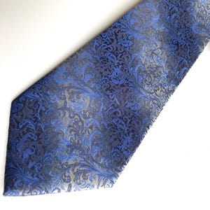 Op de afbeelding: Een blauw-grijze stropdas met een gedetailleerd bloemmotief. De stropdas is gemaakt van een glanzende stof en wordt gepresenteerd op een witte achtergrond. Het is een formeel accessoire.
