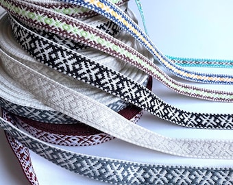 May include: A stack of woven fabric ribbons featuring intricate geometric patterns in various colors including turquoise, yellow, green, black, beige, gray, and burgundy. Each ribbon is coiled into a roll, displaying a repeating diamond design.