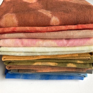 Op de afbeelding: Een stapel van acht stofvierkanten in verschillende tinten bruin, roze, geel en blauw. De stof is netjes gevouwen en heeft een zachte, textuur.