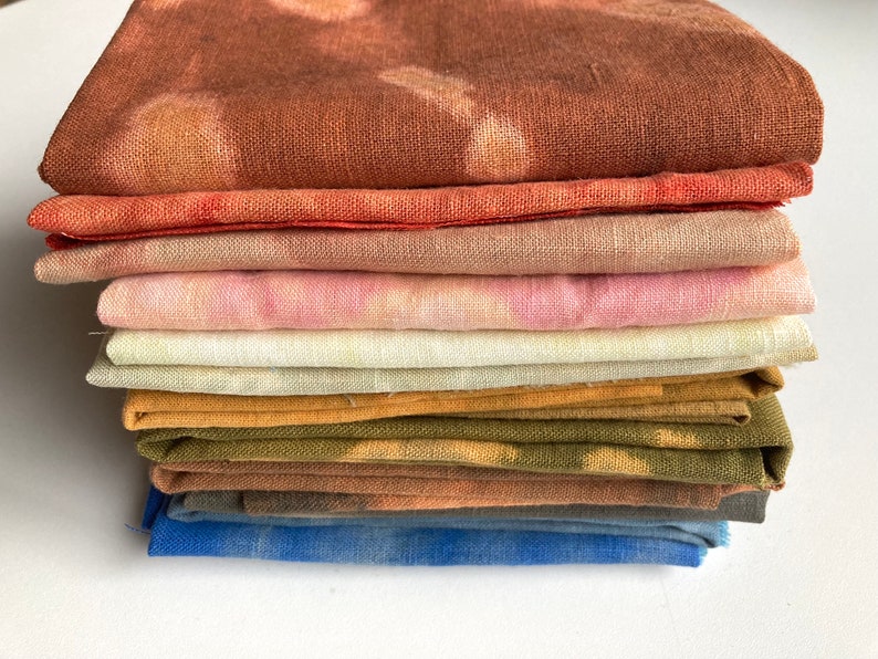 Op de afbeelding: Een stapel van acht stofvierkanten in verschillende tinten bruin, roze, geel en blauw. De stof is netjes gevouwen en heeft een zachte, textuur.