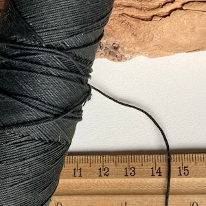 Op de afbeelding: Een spoel zwart katoenen koord met een liniaal die 11 tot 15 inch aangeeft. Het koord is ongeveer 33 cm lang.